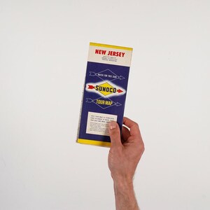 May include: A vintage Sunoco Tour-Map for New Jersey, featuring a yellow, blue, and red color scheme. The map is held in a hand against a white background. The text "NEW JERSEY" and "SUNOCO TOUR-MAP" are visible.