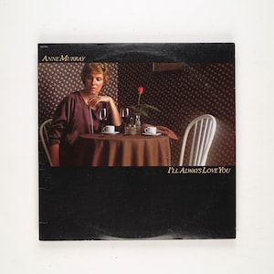 Puede incluir: Portada del álbum con Anne Murray. Está sentada a una mesa con una copa de vino y una taza de café. Una rosa roja está en un jarrón. El título del álbum "I'll Always Love You" es visible.
