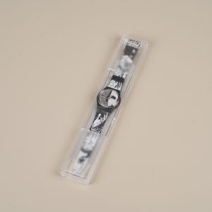 May include: A black and white Swatch watch in its original clear plastic case. The watch face and band feature a marbled design. The watch is a classic style, with a round face and a black bezel.