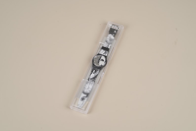 May include: A black and white Swatch watch in its original clear plastic case. The watch face and band feature a marbled design. The watch is a classic style, with a round face and a black bezel.