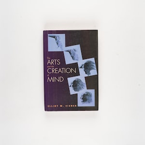 Puede incluir: Un libro de tapa dura titulado "The Arts and the Creation of Mind" de Elliot W. Eisner. El libro tiene una cubierta morada oscura con texto blanco y varias imágenes cuadradas de perfiles humanos.