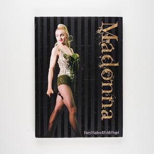 May include: A black and white striped book cover with the word "Madonna" in gold lettering. The cover features a photo of Madonna in a black and white striped outfit with a green fringe skirt. The book title is "Madonna" by Daryl Easlea & Eddi Fiegel.