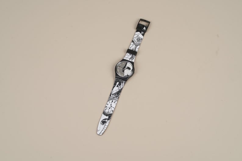 May include: A black and white wristwatch with a patterned band featuring faces and clocks. The watch has a black face and a black buckle. The band is approximately 20 cm long.