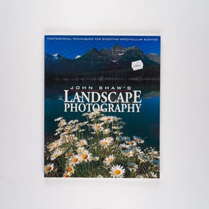 May include: A book titled "John Shaw's Landscape Photography: Professional Techniques for Shooting Spectacular Scenes" with a photo of a mountain lake and wildflowers on the cover.