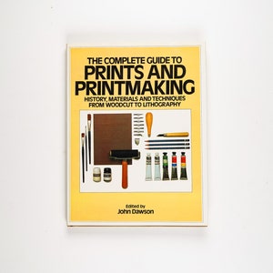 Puede incluir: La portada del libro "La guía completa de grabados y grabado" con un fondo amarillo y una variedad de herramientas y materiales de grabado, incluyendo pinceles, lápices, tubos de pintura y un rodillo. El texto de la portada dice "La guía completa de grabados y grabado" y "Historia, materiales y técnicas del grabado en madera a la litografía".