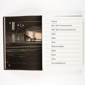 May include: A black and white photograph of an open book. The left page features a close-up of a record player. The right page displays a table of contents with the headings: Preface, 1857-1919: The Acoustic Era, and other decades.