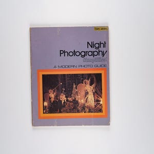 Könnte beinhalten: Ein lilafarbenes Buch mit dem Titel "Night Photography Simplified" mit einem modernen Fotoguide. Der Umschlag zeigt ein Foto einer Nachtszene mit Lichtern und einer Person in einem Kostüm.
