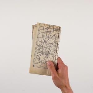May include: A stack of vintage maps, featuring a detailed road network in black ink on aged, cream-colored paper. The maps are held in a hand against a plain white background, showcasing the aged texture and intricate details of the cartography.