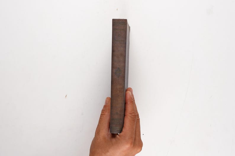 May include: A brown, rectangular book is held vertically against a white background. The book's spine is visible, showing a slightly worn, textured surface. The book is held by a hand.