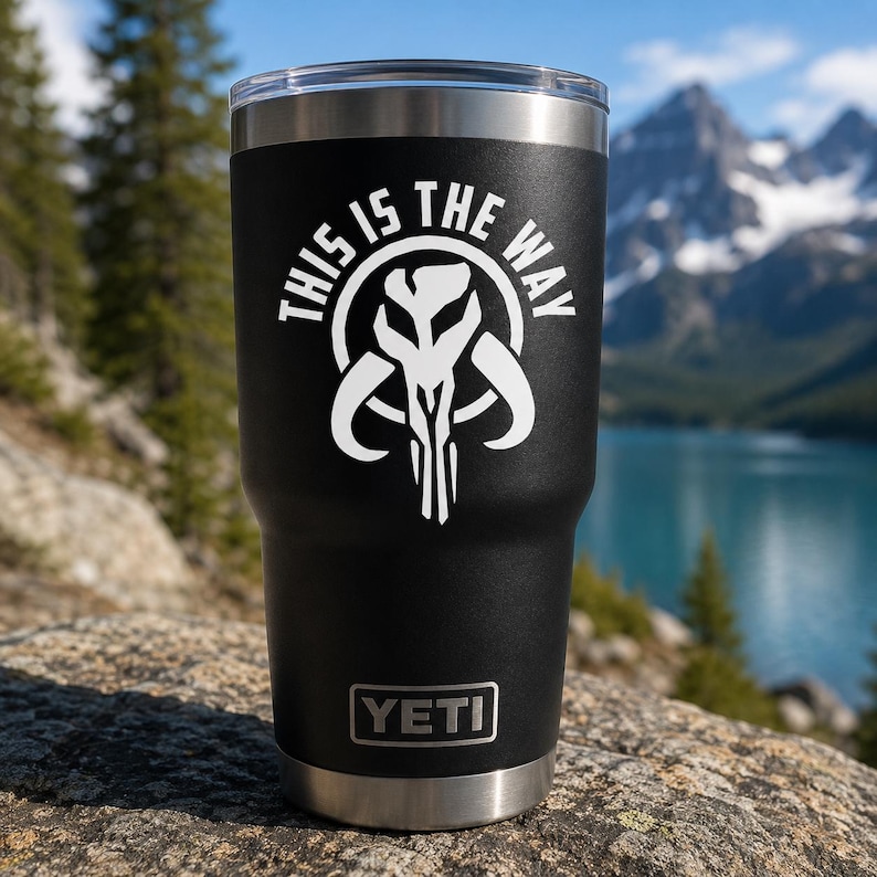May include: A black stainless steel tumbler featuring a white graphic of a Mythosaur skull emblem and the text "THIS IS THE WAY" arched above it. The base of the tumbler displays the white YETI brand logo on a metallic silver rim.