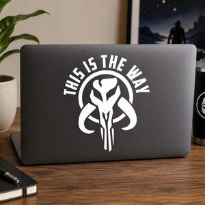 May include: A dark gray laptop featuring a white decal of a stylized skull emblem centered on the lid. The decal includes the text "THIS IS THE WAY" arched over the top of the skull graphic in a bold, sans-serif font, creating a high-contrast design.
