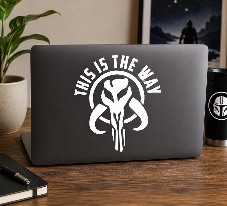 May include: A dark gray laptop featuring a white decal of a stylized skull emblem centered on the lid. The decal includes the text "THIS IS THE WAY" arched over the top of the skull graphic in a bold, sans-serif font, creating a high-contrast design.