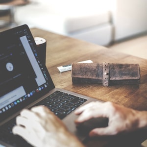 Pode incluir: Uma pessoa está trabalhando em um laptop com um estojo de lápis de couro marrom em uma mesa de madeira.