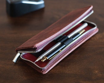 May include: A brown leather pencil case with a zipper closure. The case is open and contains two silver and black pens.