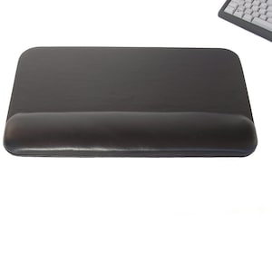 Puede incluir: Alfombrilla de ratón rectangular negra con reposamuñecas integrado. La alfombrilla está hecha de un material texturizado y tiene esquinas redondeadas. El reposamuñecas es acolchado y curvado para soporte. Un teclado es visible en el fondo.