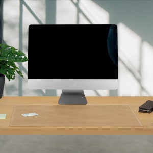 May include: A light brown leather desk mat with a stitched edge. The mat is on a wooden desk with a computer monitor and a small black wallet on the surface.