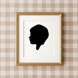 May include: A framed silhouette portrait of a person in profile, rendered in black against a white background. The portrait is in a gold frame, with the word "sister" written in cursive below the silhouette. The background is a tan and white checkered pattern.