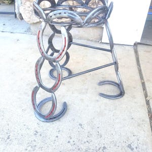 May include: A plant stand made from horseshoes. The stand has a circular top and three legs. The horseshoes are welded together and have a red stripe painted on them.
