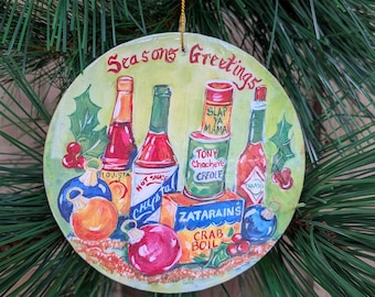 May include: Round Christmas ornament with the words "Seasons Greetings". The ornament features illustrations of hot sauce bottles, a can of "Slap Ya Mama" seasoning, and a box of Zatarain's Crab Boil. Colorful baubles and holly complete the design.