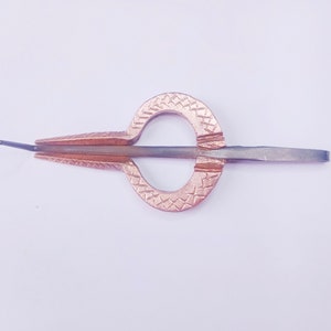 May include: A bronze-coloured jaw harp, also known as a mouth harp, on a white background. The instrument has a rounded, patterned frame and a central metal reed. The tines are angled, and the instrument is designed to be held in the mouth.