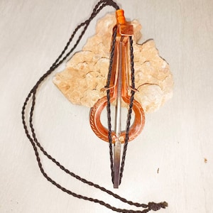 Op de afbeelding: Een koperkleurige mondharp, ook wel mondharp genoemd, met een donkerbruin touw. Het instrument rust op een lichtbruine, getextureerde steen. De harp heeft een cirkelvormig resonerend lichaam en een metalen tong.