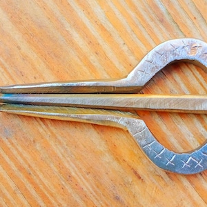 May include: A silver jaw harp, a musical instrument, is displayed on a wooden surface. The harp features a curved frame with etched patterns and a thin, curved metal tongue. The instrument is designed to be held in the mouth and plucked.