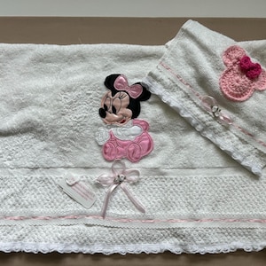 May include: White baby towel set with a Minnie Mouse applique in pink and black. The set includes a large towel, a smaller hooded towel, and decorative bows. The towels have lace trim and pink accents.