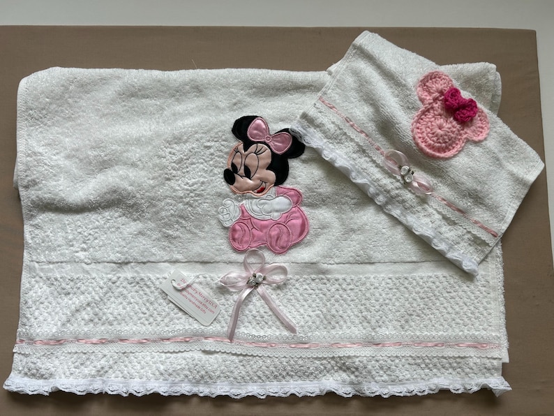 May include: White baby towel set with a Minnie Mouse applique in pink and black. The set includes a large towel, a smaller hooded towel, and decorative bows. The towels have lace trim and pink accents.