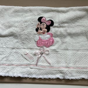 May include: White baby towel featuring a Minnie Mouse applique in pink and black. The towel has a decorative lace trim with a pink ribbon and bow. A small tag is attached.