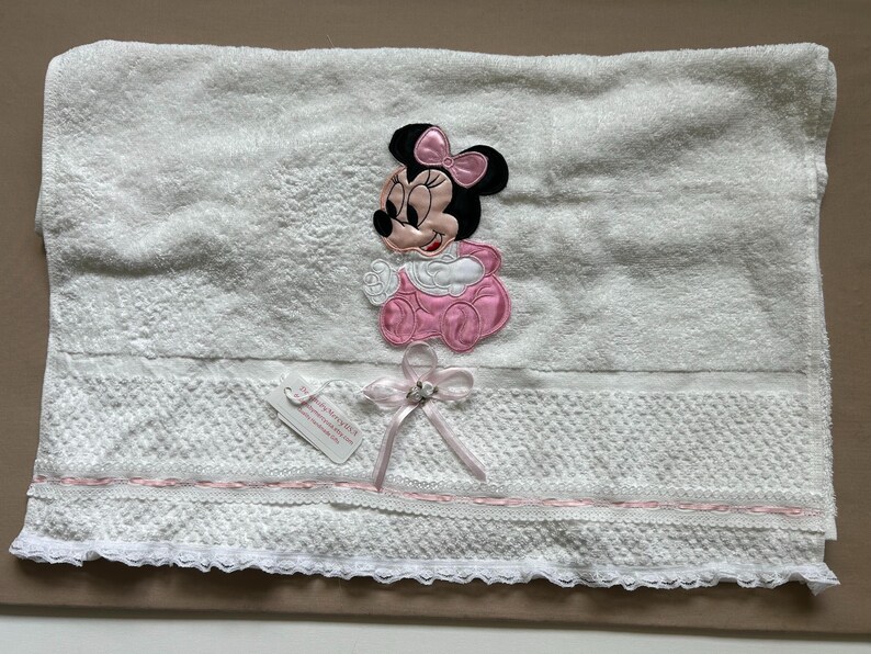 May include: White baby towel featuring a Minnie Mouse applique in pink and black. The towel has a decorative lace trim with a pink ribbon and bow. A small tag is attached.