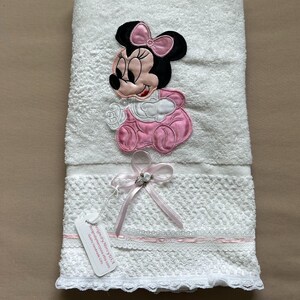 May include: White baby towel with an embroidered Minnie Mouse design in pink, black, and white. The towel has a decorative lace trim and a pink ribbon with a small embellishment. A tag is attached to the towel.