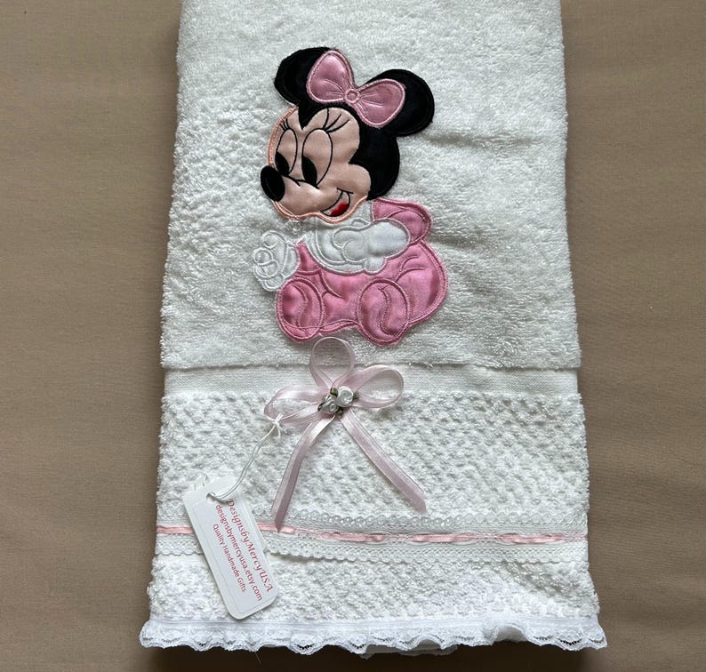 May include: White baby towel with an embroidered Minnie Mouse design in pink, black, and white. The towel has a decorative lace trim and a pink ribbon with a small embellishment. A tag is attached to the towel.