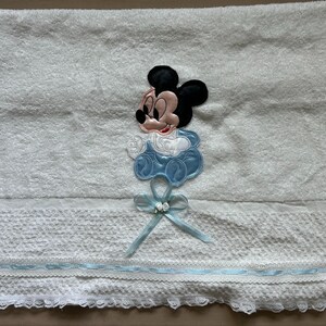 Puede incluir: Toalla de beb&eacute; blanca con una aplicaci&oacute;n de Mickey Mouse. La toalla presenta un dise&ntilde;o de Mickey Mouse azul y blanco, una cinta azul y un borde de encaje decorativo. La toalla est&aacute; hecha de material suave y absorbente.