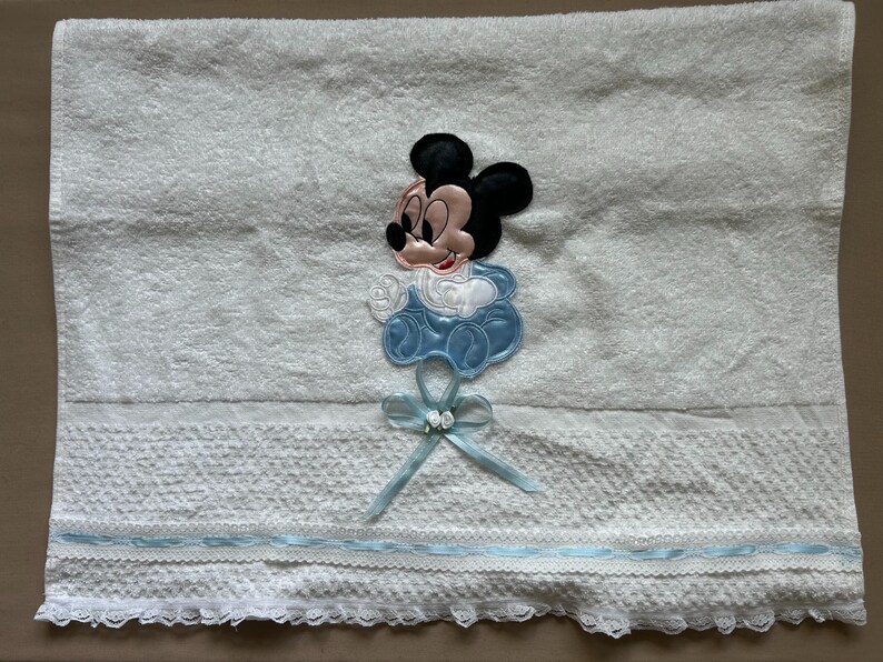 Puede incluir: Toalla de beb&eacute; blanca con una aplicaci&oacute;n de Mickey Mouse. La toalla presenta un dise&ntilde;o de Mickey Mouse azul y blanco, una cinta azul y un borde de encaje decorativo. La toalla est&aacute; hecha de material suave y absorbente.