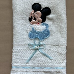 Puede incluir: Toalla de beb&eacute; blanca con un dise&ntilde;o bordado de Mickey Mouse. La toalla presenta un lazo azul con flores blancas y un ribete azul. El dise&ntilde;o de Mickey Mouse es en negro, rosa y azul.