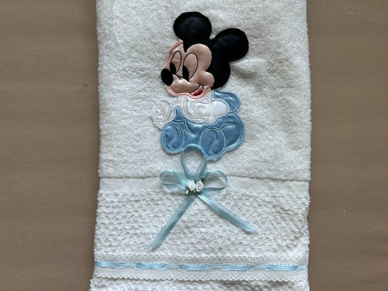 Puede incluir: Toalla de beb&eacute; blanca con un dise&ntilde;o bordado de Mickey Mouse. La toalla presenta un lazo azul con flores blancas y un ribete azul. El dise&ntilde;o de Mickey Mouse es en negro, rosa y azul.