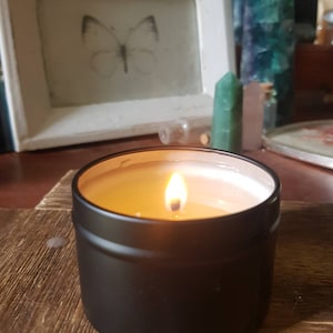 May include: A black candle tin with a lit candle inside. The candle is a light yellow color and the wax is smooth. The tin is sitting on a wooden surface.