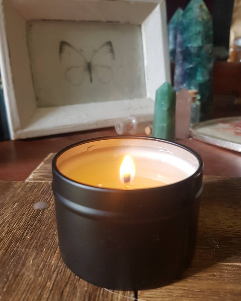 May include: A black candle tin with a lit candle inside. The candle is a light yellow color and the wax is smooth. The tin is sitting on a wooden surface.