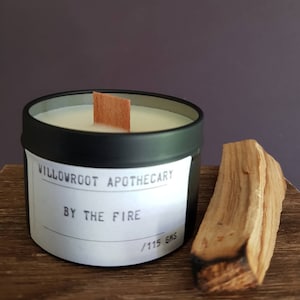 May include: Black candle tin with a white label that reads "Willowroot Apothecary By The Fire / 115 gms". A wooden wick sits in the candle. A piece of brown wood is in the foreground.