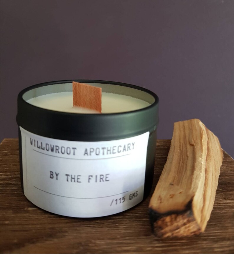 May include: Black candle tin with a white label that reads "Willowroot Apothecary By The Fire / 115 gms". A wooden wick sits in the candle. A piece of brown wood is in the foreground.