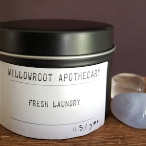 May include: A black metal tin with a white label that reads "WILLOWROOT APOTHECARY FRESH LAUNDRY" and "115 gms". The tin is next to three stones, one blue and two clear, on a wooden surface.