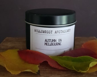 May include: A black cylindrical metal candle tin with a white label featuring the text Willowroot Apothecary and Autumn in Melbourne. The container is surrounded by several fallen leaves in shades of yellow, orange, and deep red.
