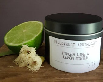 Op de afbeelding: Een zwarte blikken kaars met een wit label waarop staat "Willowroot Apothecary Finger Lime & Lemon Myrtle /15". Op de voorgrond liggen een limoenpartje en een witte bloem.