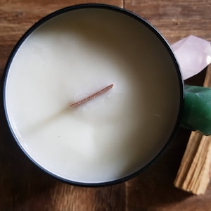 May include: A black candle jar with a white wax candle and a wooden wick. The candle is surrounded by a pink crystal, a green crystal, and a piece of palo santo wood.