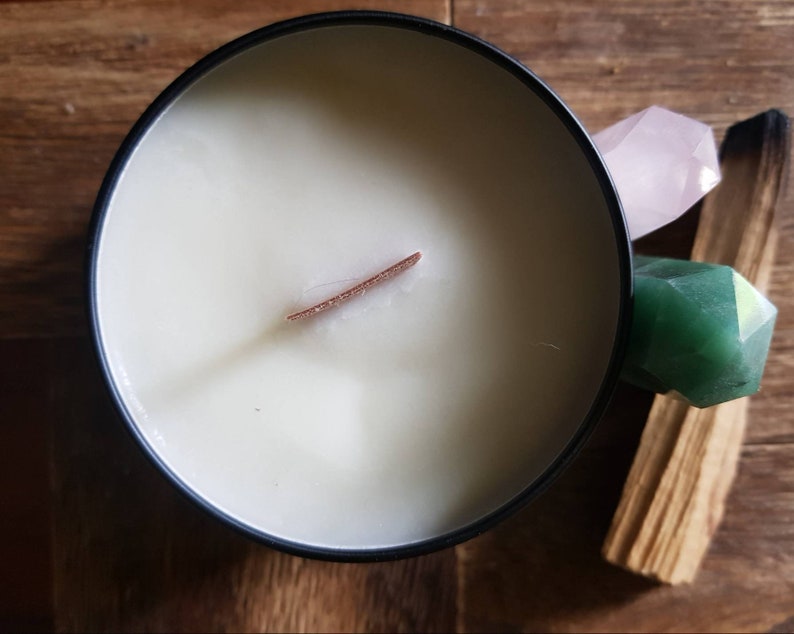 May include: A black candle jar with a white wax candle and a wooden wick. The candle is surrounded by a pink crystal, a green crystal, and a piece of palo santo wood.