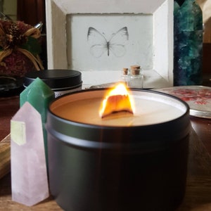 May include: A black candle in a tin container with a wooden wick burning. The candle is surrounded by crystals and a small picture frame with a butterfly illustration.
