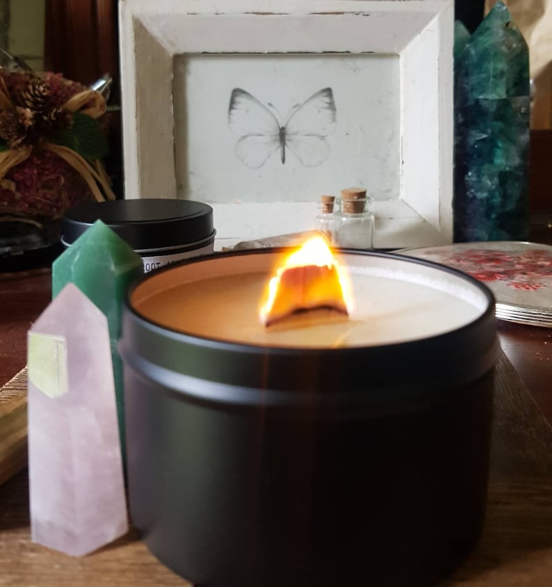 May include: A black candle in a tin container with a wooden wick burning. The candle is surrounded by crystals and a small picture frame with a butterfly illustration.