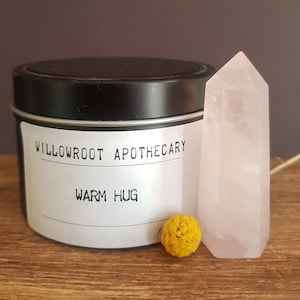 May include: A black tin candle with a white label that reads "Willowroot Apothecary Warm Hug". A pink crystal and a yellow flower are next to the candle.