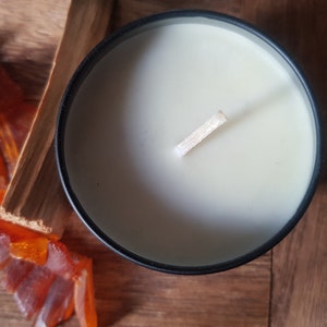 May include: A black candle jar with a white wax candle and a wooden wick. The candle is on a wooden surface with orange glass pieces.