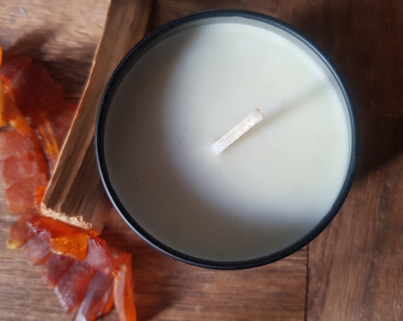 May include: A black candle jar with a white wax candle and a wooden wick. The candle is on a wooden surface with orange glass pieces.